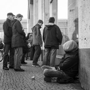 A homeless man sits on the sidewalk on a busy street as crowds of shoppers stand nearby.