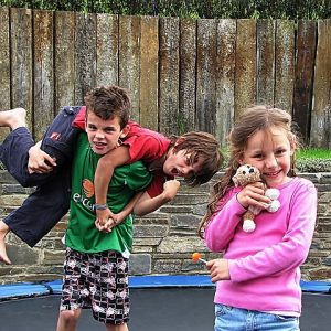 Two young boys wrestle in the yard while as a girl stands by calmly holding a stuffed animal.