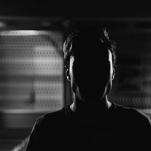 A man stands in a darkened room with his face almost entirely obscured by shadow.