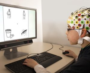 A boy wearing an EEG cap uses a mouse and keyboard while looking at images on a computer monitor during an experiment.