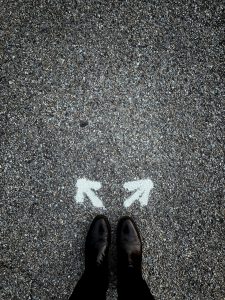 Pavement. On the bottom are two feet in black boots pointed at two arrows. One arrow points to forward to the left and the other arrow points to the forward and right. The image indicates that the person much choose a direction to go forward.
