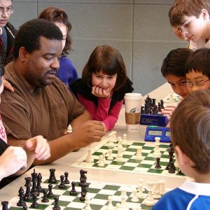 A group of children watch as adults play a game of chess.