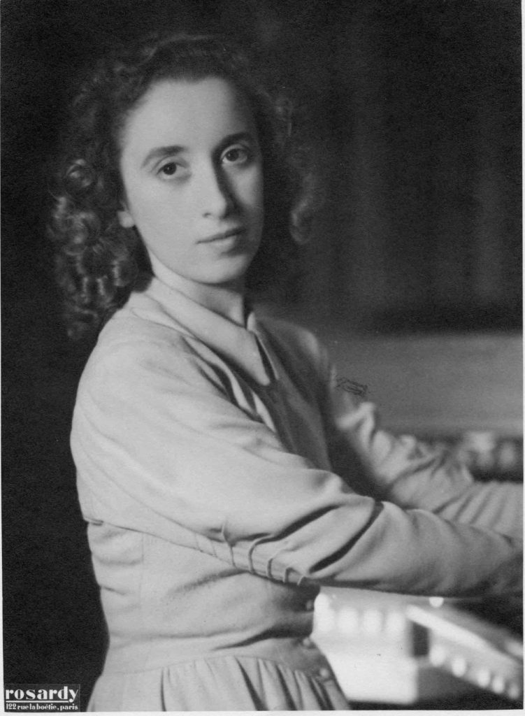 Head-torso-and-forearms view, slightly angled, of a young woman, wearing long ringlets and a serious expression on her face, seated at an organ console, with her head turned to look at the photographer.
