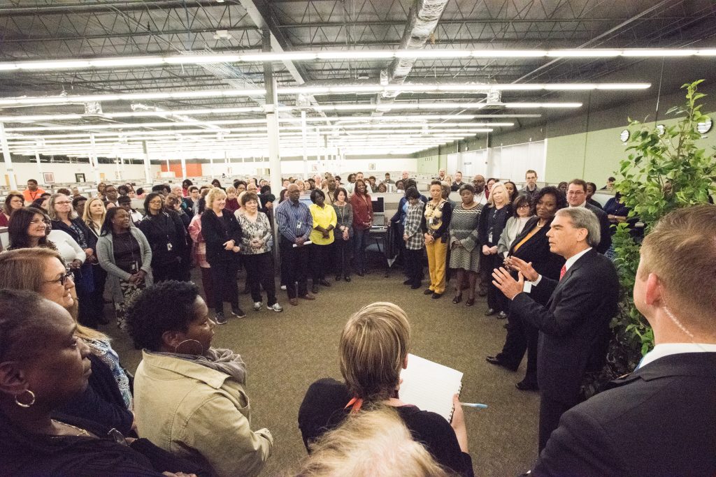 A large group of workers in an open concept office gather around a man in a suit who is explaining something.