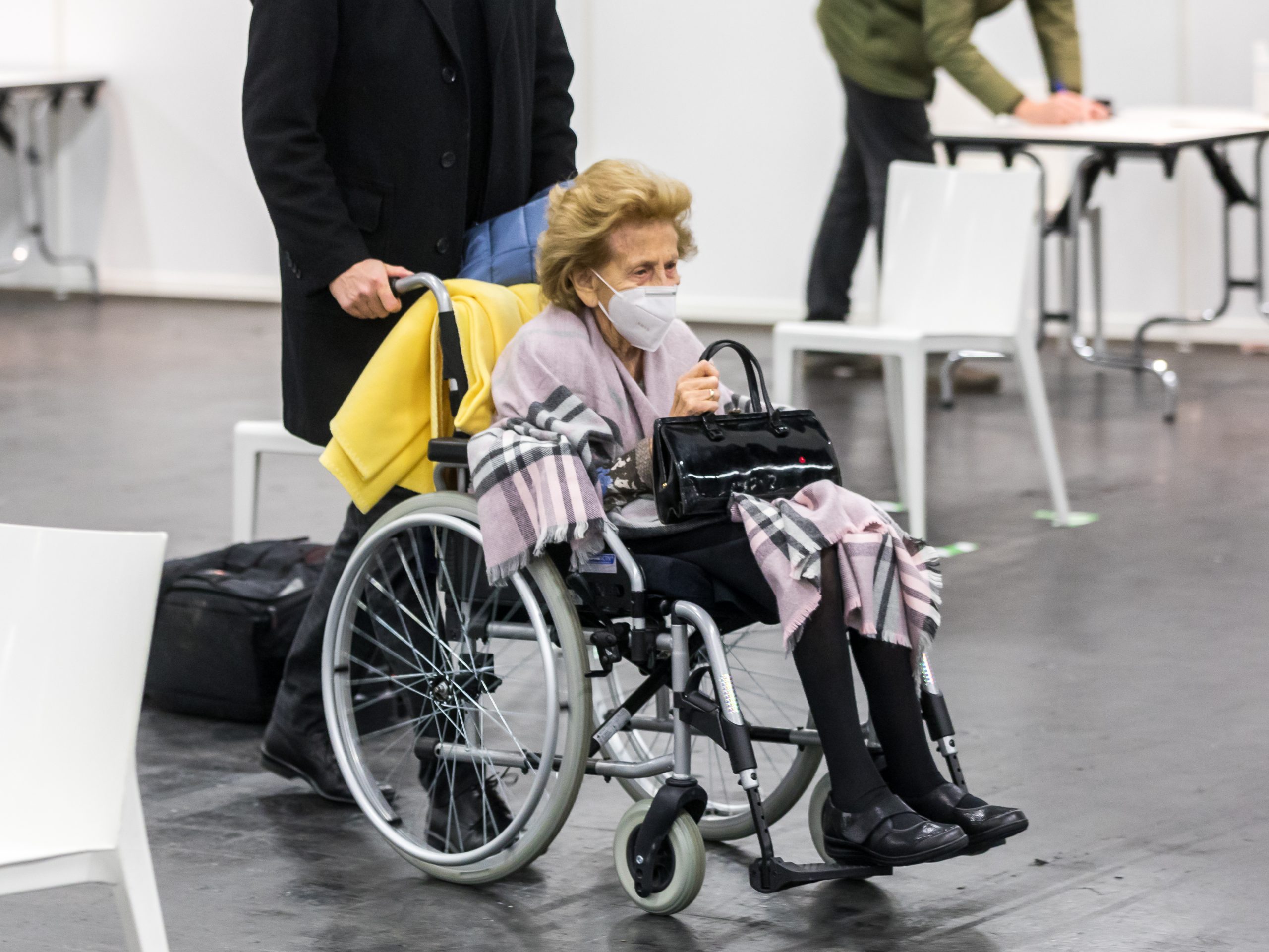 ”COVID-19_vaccination_center,_fair_grounds_Cologne,_1st_vaccination-8070.jpg” by Raimond Spekking is licensed under CC BY-SA 4.0 Photo showing an elderly woman, wearing a mask being pushed in a wheelchair
