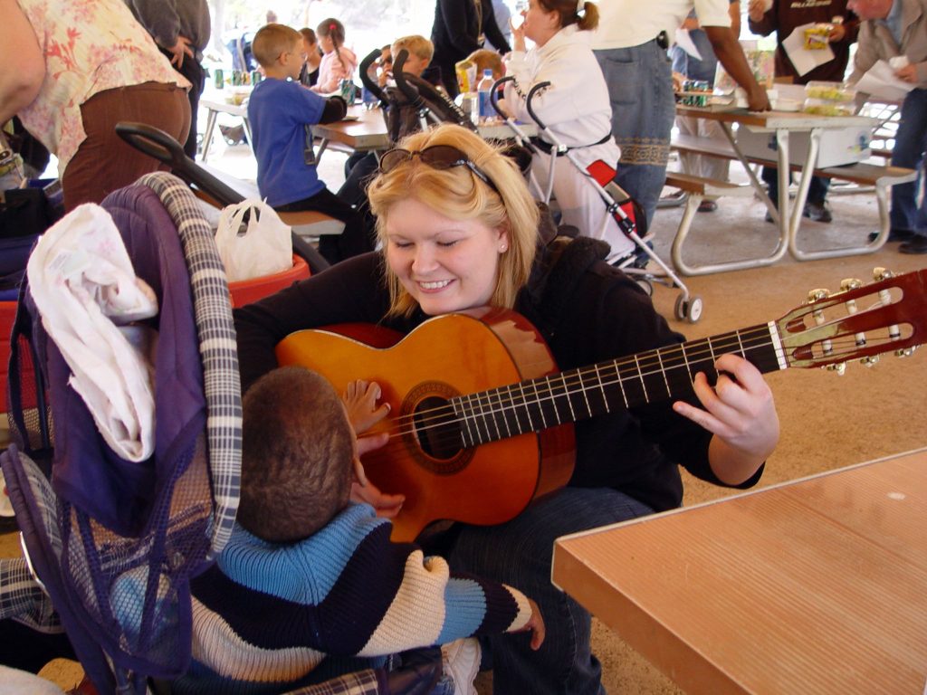 ”103909417_f36b60ceec_k” by midiman is licensed under CC BY 2.0 Photo of a woman holding a guitar, smiling at a child who is touching the guitar. Other people are shown in the background.